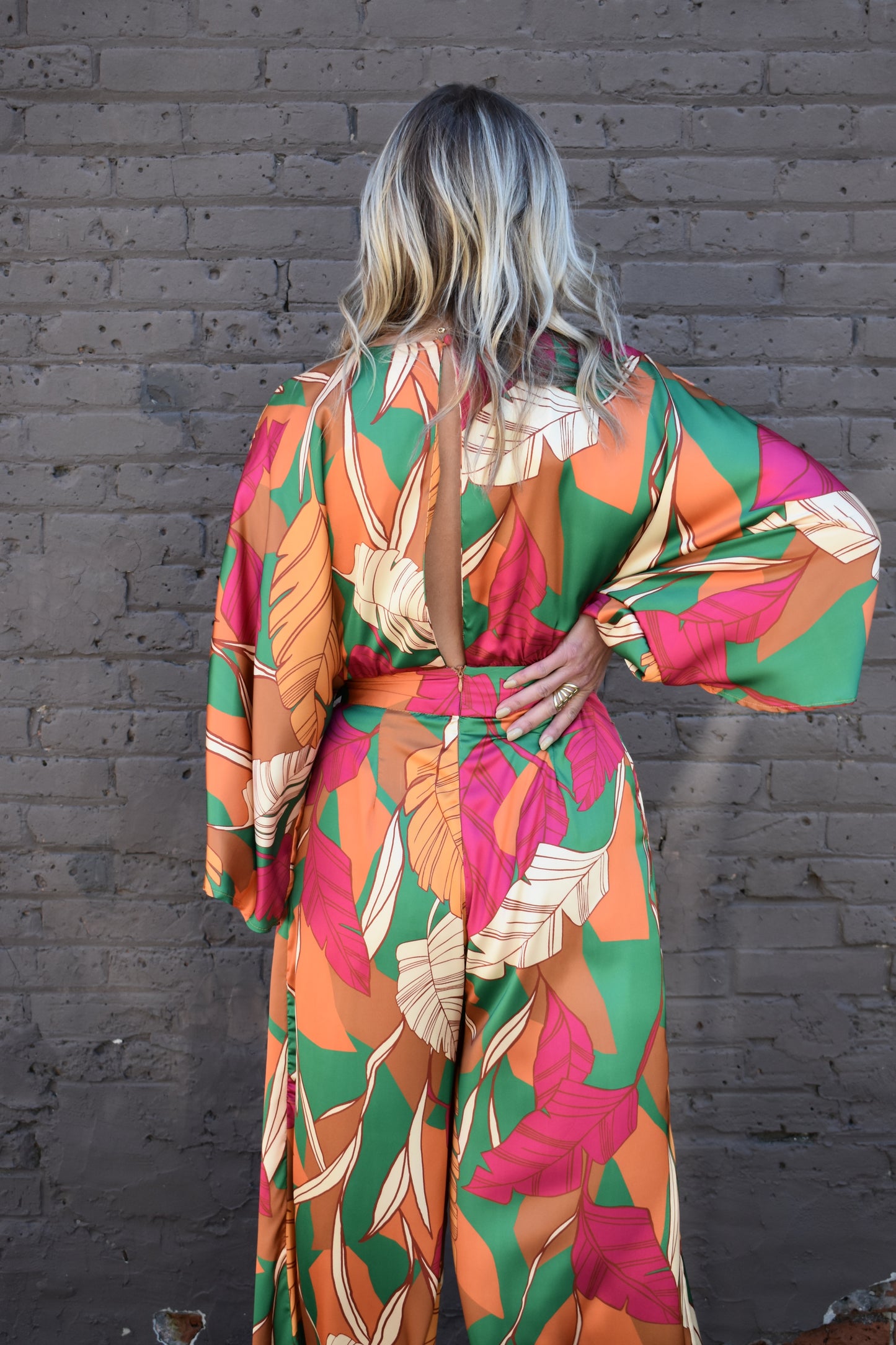statement satin jumpsuit with a green, rust, cream and pink color scheme. V neckline, kimono sleeves, wide waistband, side pockets, center back slit with button closure on back of neck. wide leg.