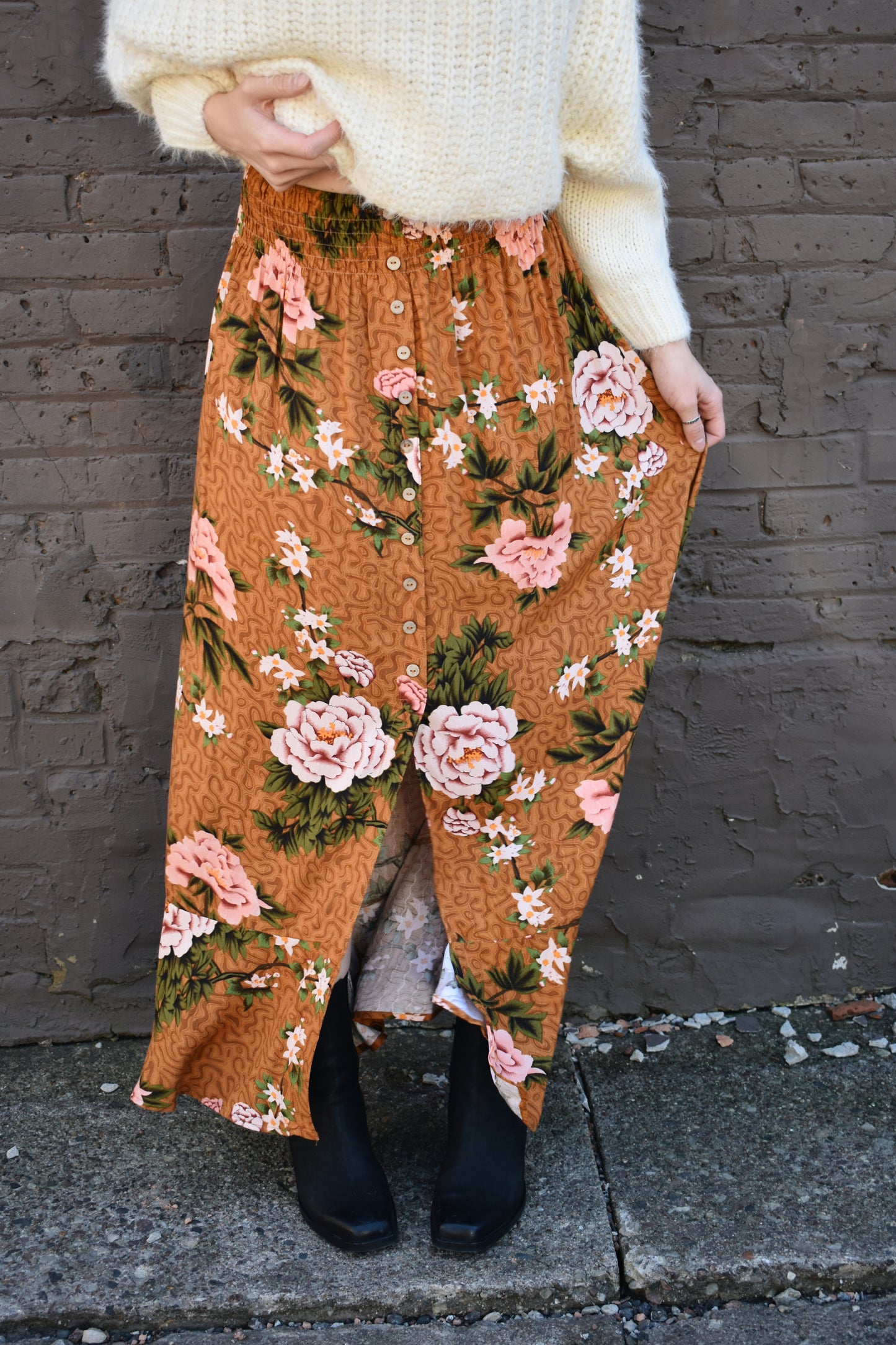 Floral printed maxi skirt with a 3/4 button down front that leads into a center slit. slight ruffle at the bottom. elastic waistband. light brown/rust color background with big printed white and blush colored flowers with green stems.