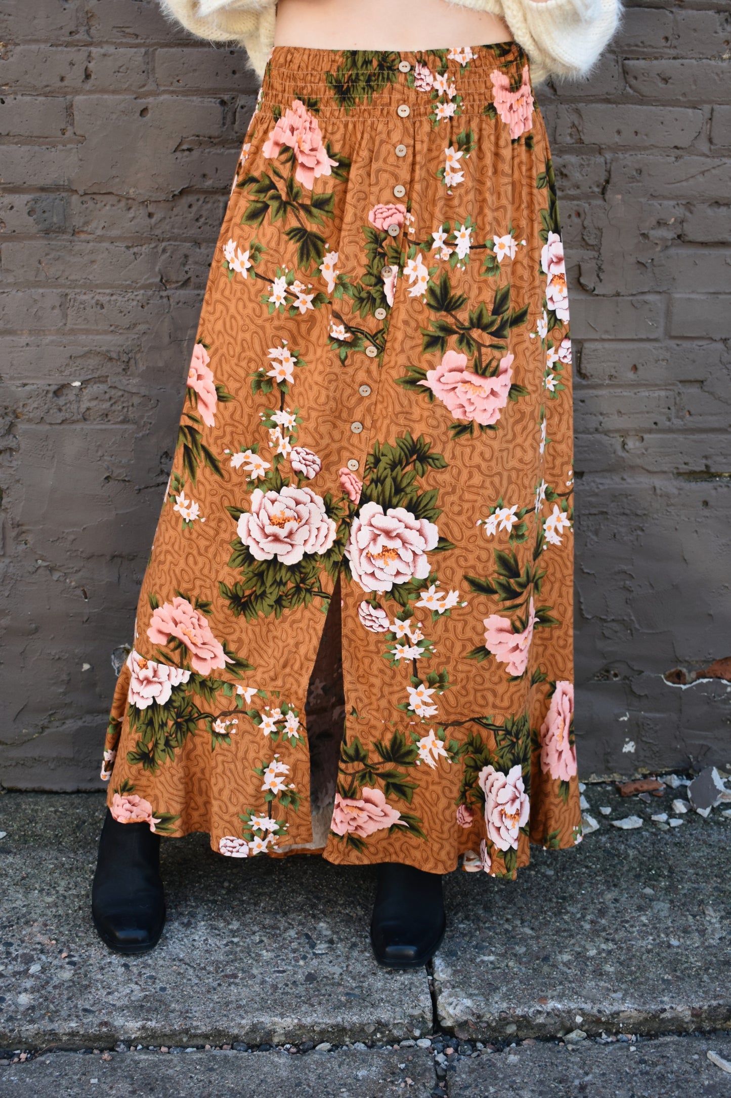 Floral printed maxi skirt with a 3/4 button down front that leads into a center slit. slight ruffle at the bottom. elastic waistband. light brown/rust color background with big printed white and blush colored flowers with green stems.