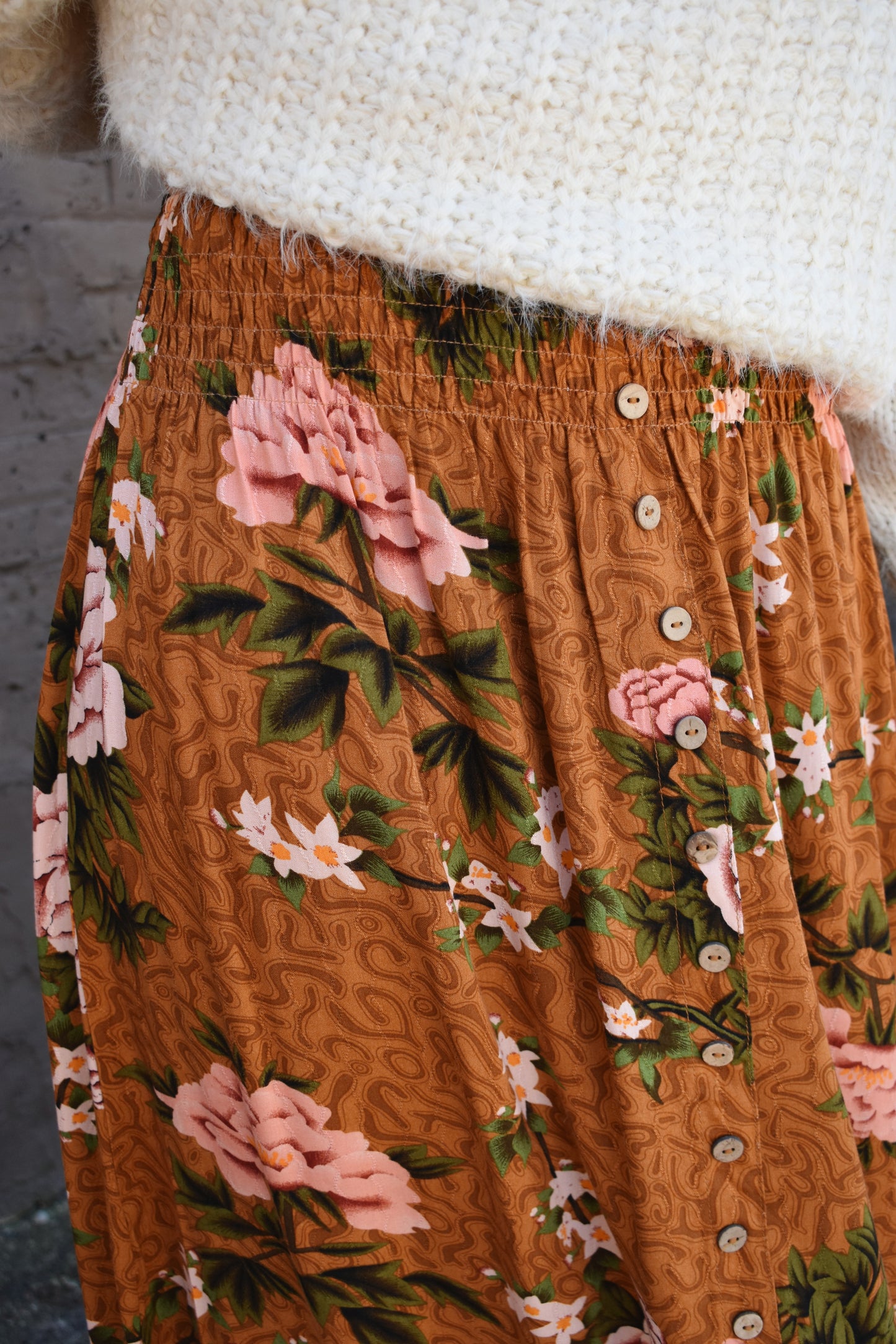 Floral printed maxi skirt with a 3/4 button down front that leads into a center slit. slight ruffle at the bottom. elastic waistband. light brown/rust color background with big printed white and blush colored flowers with green stems.