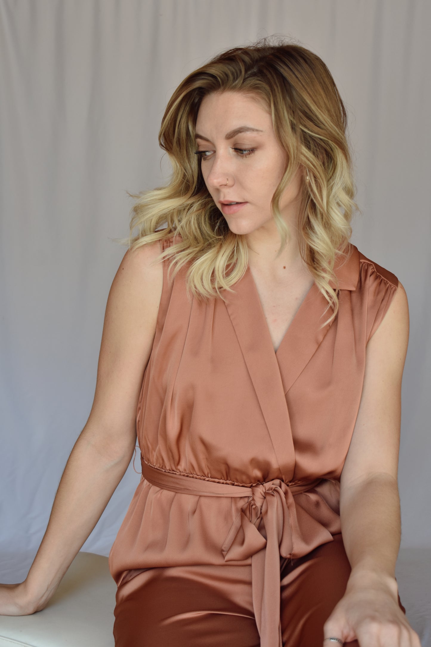 collared v neck sleeveless satin copper toned jumpsuit. Elastic waist with sash tie. front pockets. rushing on top of shoulders.