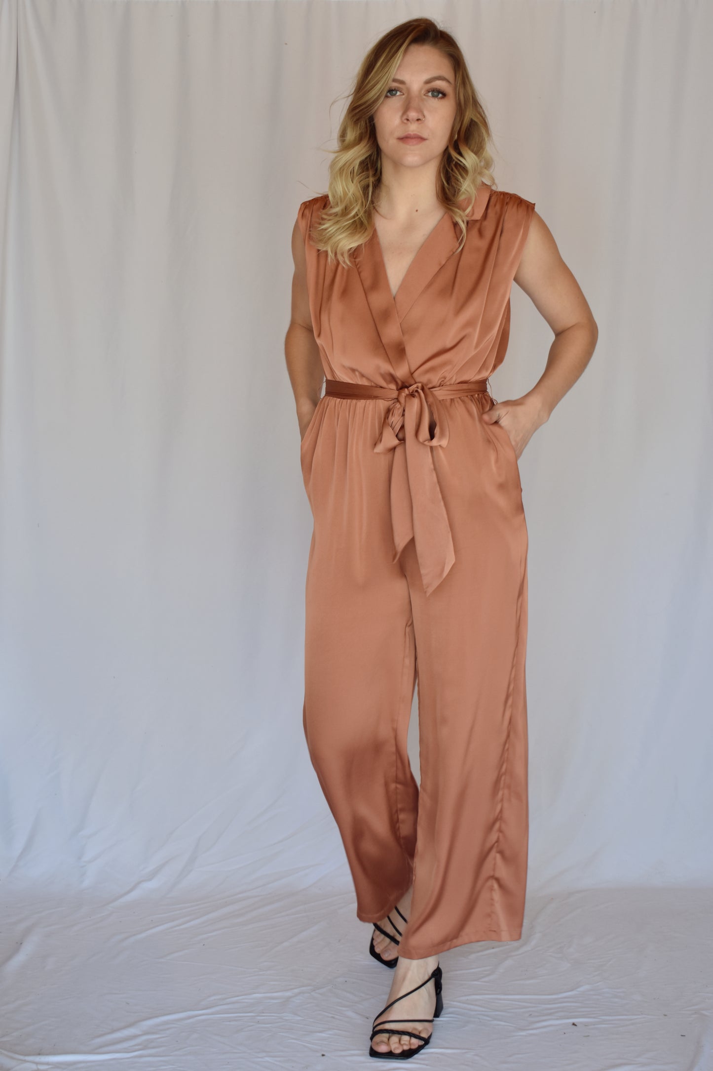 collared v neck sleeveless satin copper toned jumpsuit. Elastic waist with sash tie. front pockets. rushing on top of shoulders.