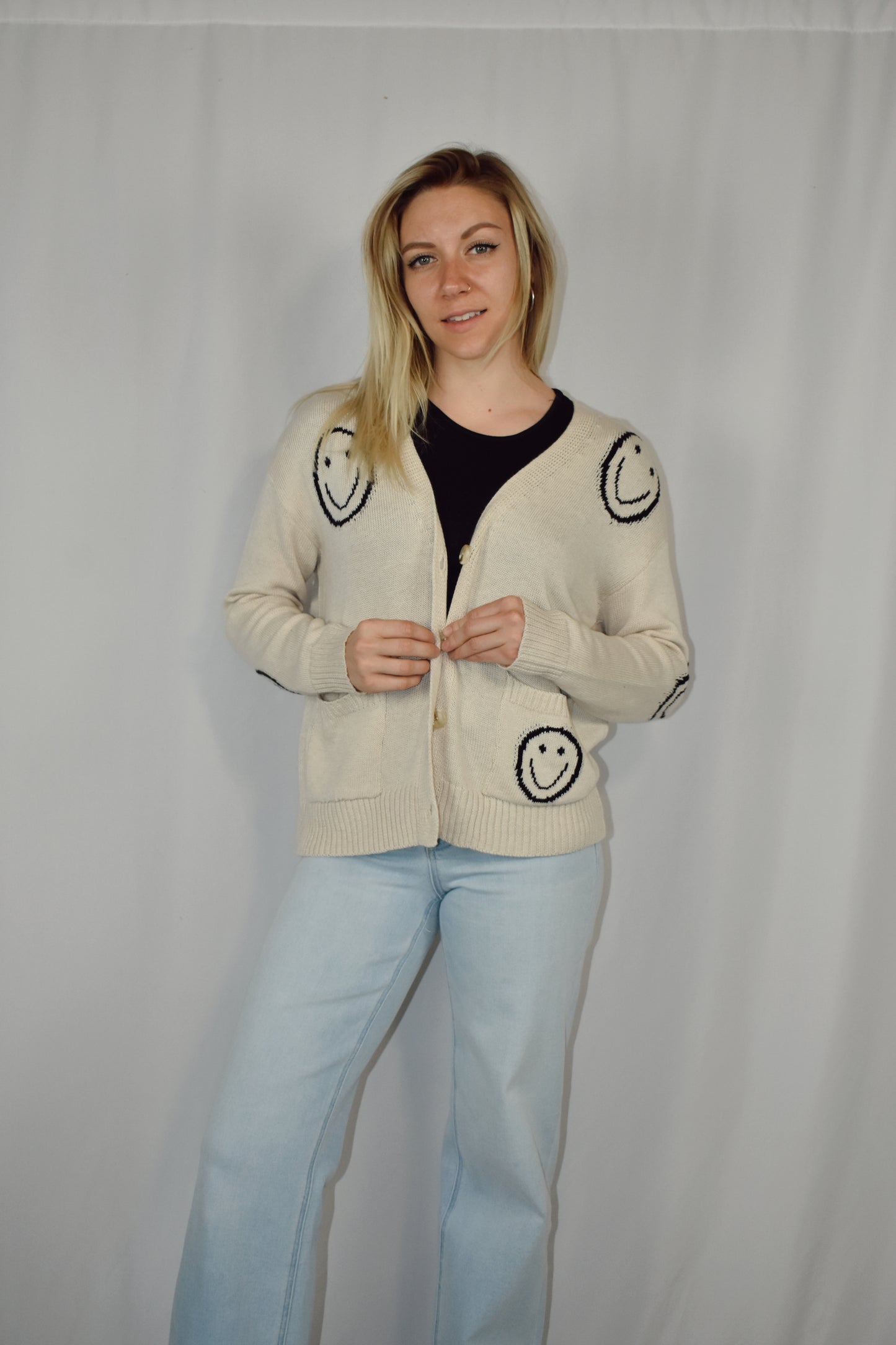 Button down cardigan in a neutral color with big black smiley faces and two patch pockets on the front. Waist length