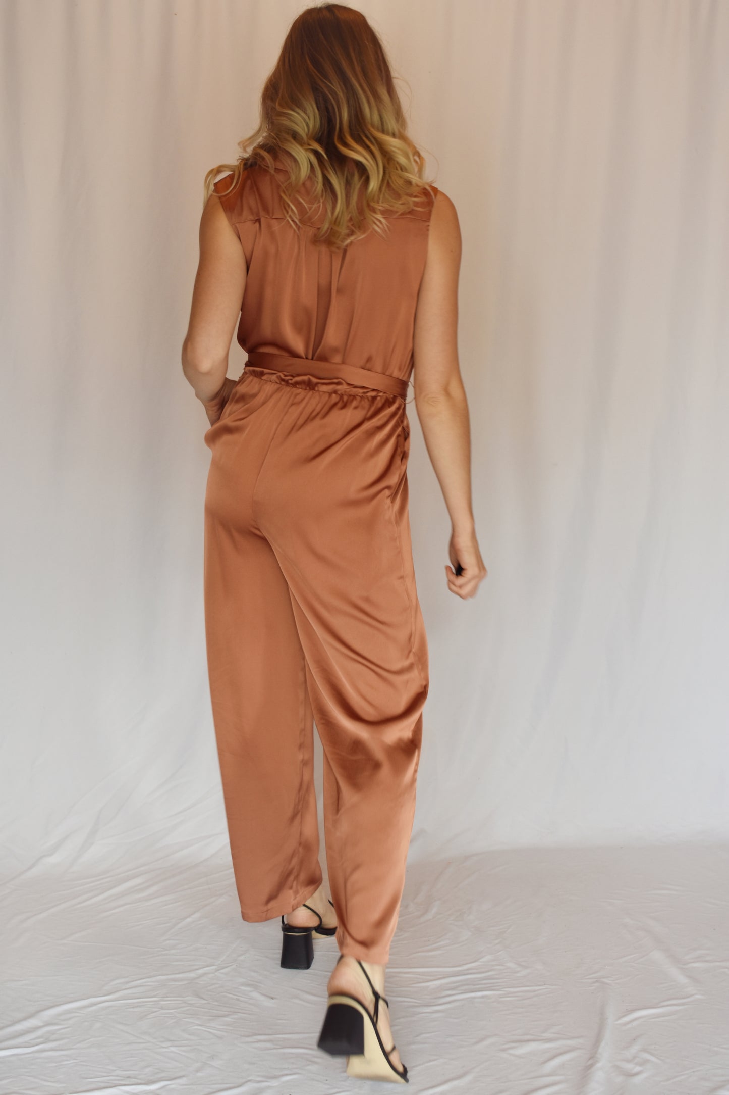 collared v neck sleeveless satin copper toned jumpsuit. Elastic waist with sash tie. front pockets. rushing on top of shoulders.