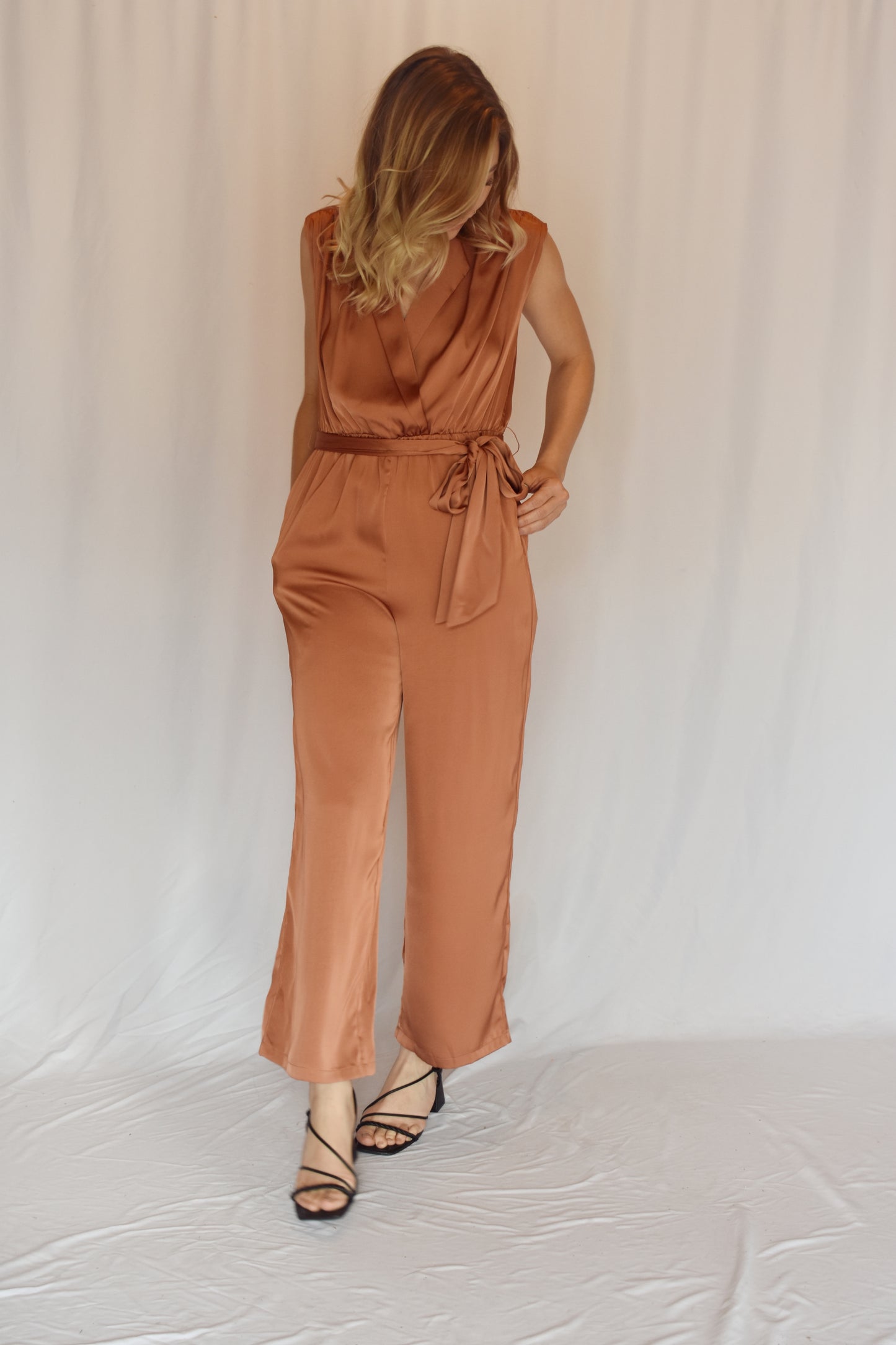 collared v neck sleeveless satin copper toned jumpsuit. Elastic waist with sash tie. front pockets. rushing on top of shoulders.