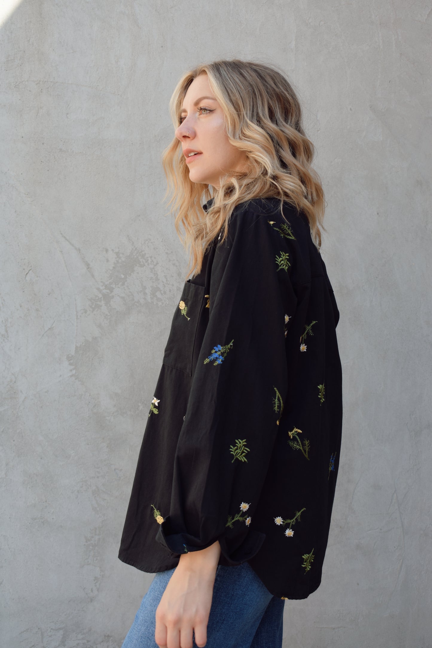 oversized black button down with flowers embroidered on front, back, and sleeves. Flowers are green, blue, yellow, white. Shirt tail hem, buttoned cuffs, one patch breast pockets, collared neckline, black buttons.