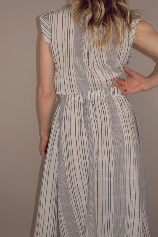 white button down midi dress with blue vertical stripes. cap short sleeves with cuff and button on top of the shoulder. skirt has extra fabric that comes together and ties in the front making skirt more full and flowy.  back has synching at waist with cut out that meets waist band.