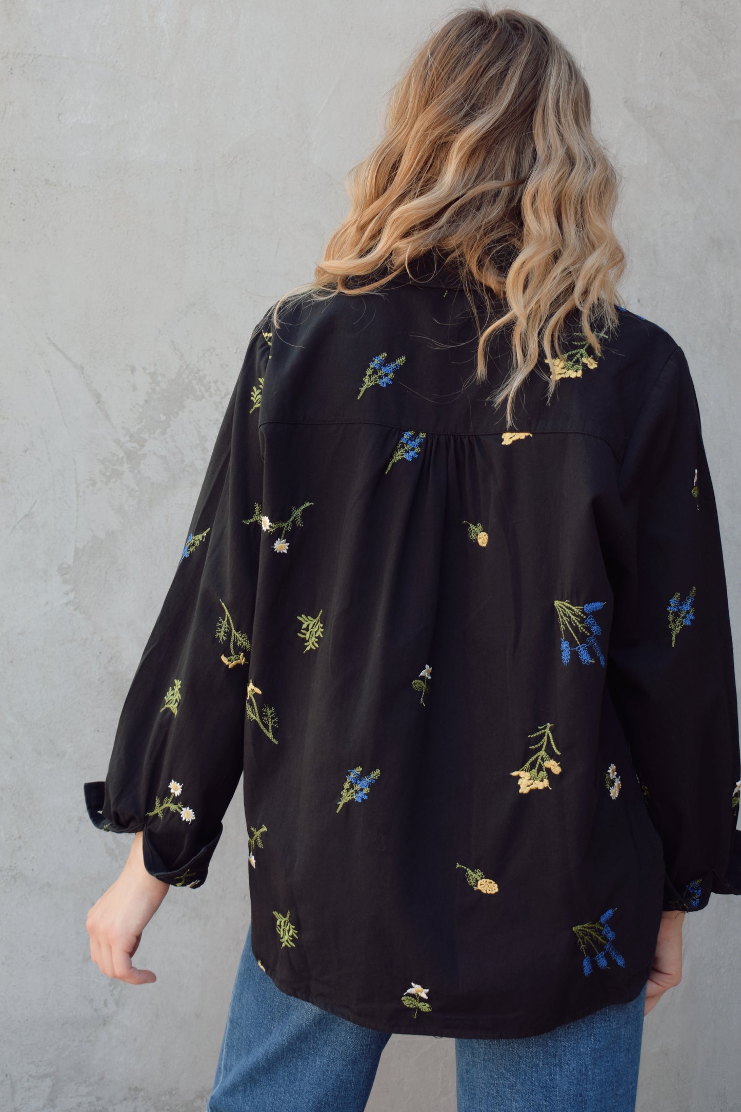 oversized black button down with flowers embroidered on front, back, and sleeves. Flowers are green, blue, yellow, white. Shirt tail hem, buttoned cuffs, one patch breast pockets, collared neckline, black buttons.