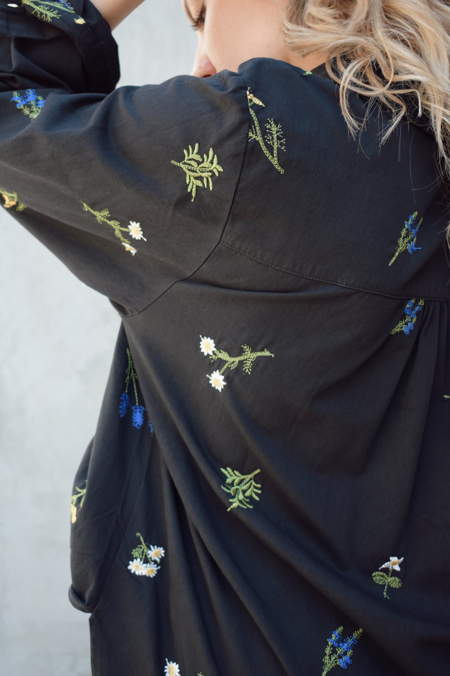 oversized black button down with flowers embroidered on front, back, and sleeves. Flowers are green, blue, yellow, white. Shirt tail hem, buttoned cuffs, one patch breast pockets, collared neckline, black buttons.