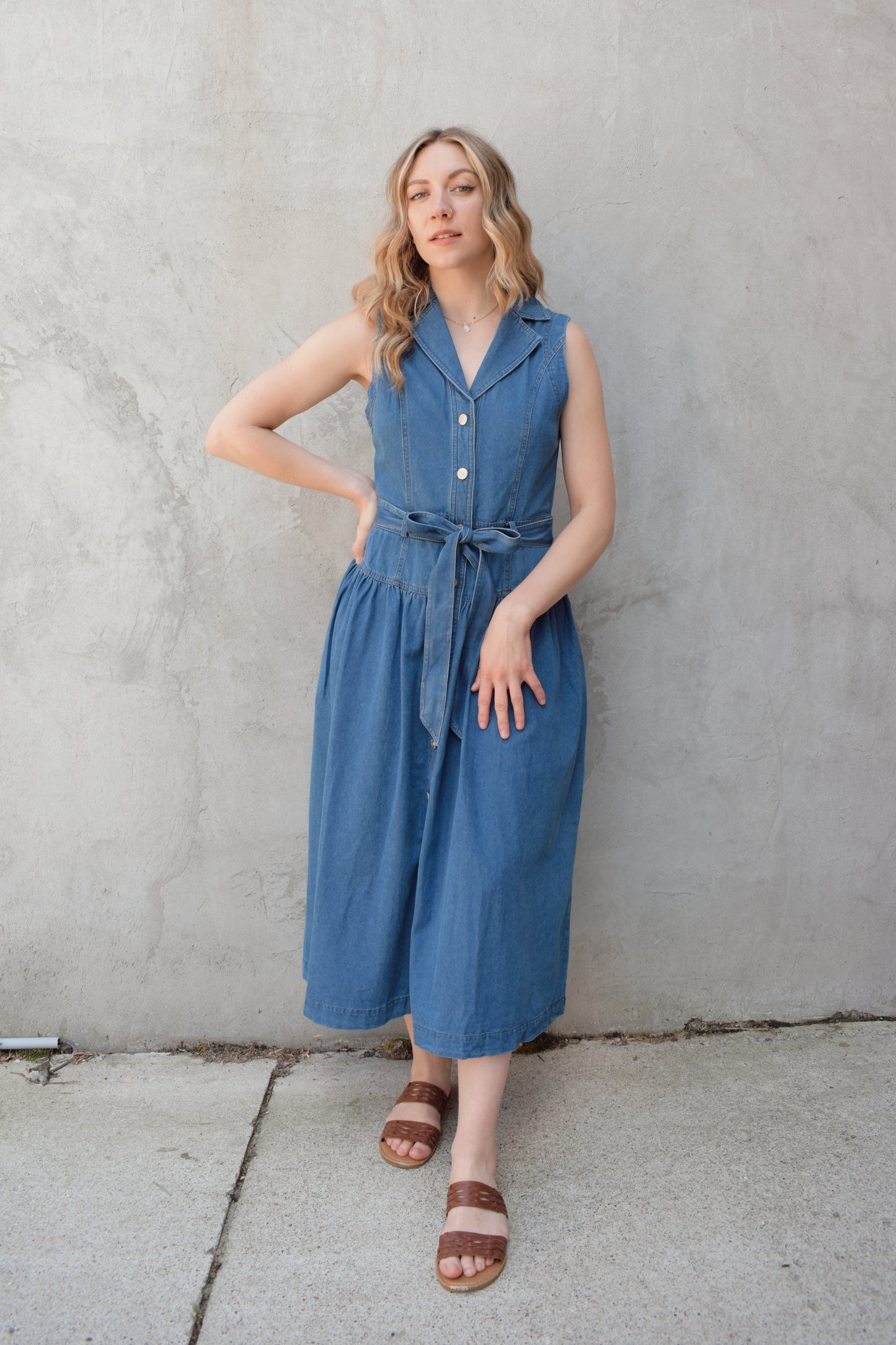 sleeveless medium wash denim midi dress with notched lapel collar, buttons down the entire front, drop waist, beltloops with denim tie at waist, has pockets