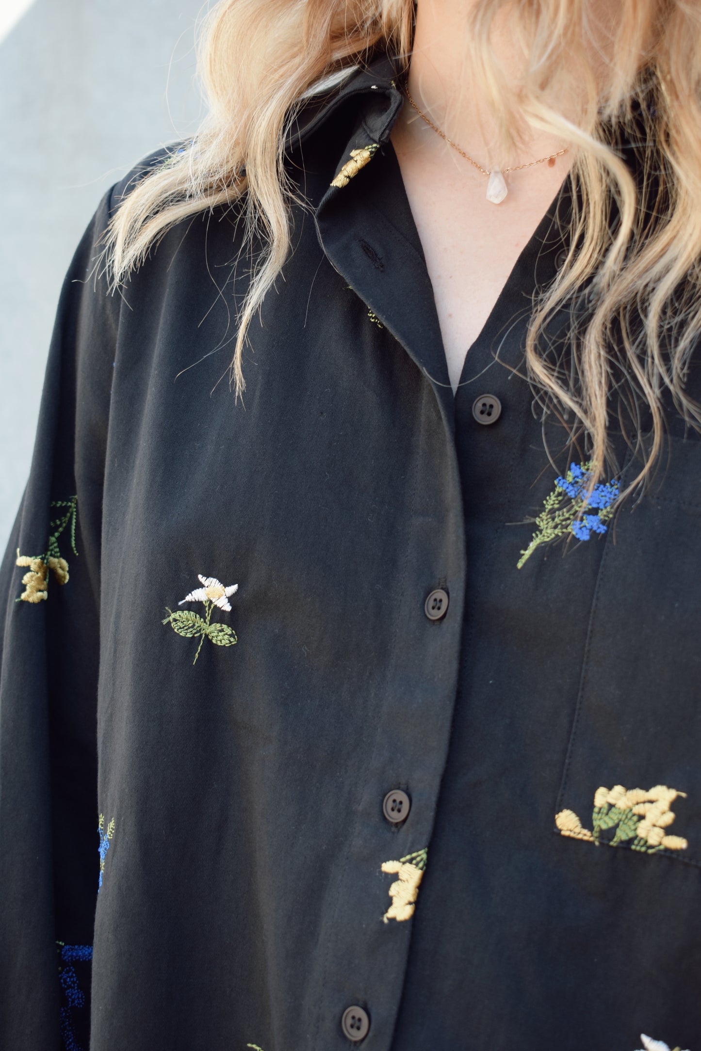 oversized black button down with flowers embroidered on front, back, and sleeves. Flowers are green, blue, yellow, white. Shirt tail hem, buttoned cuffs, one patch breast pockets, collared neckline, black buttons.