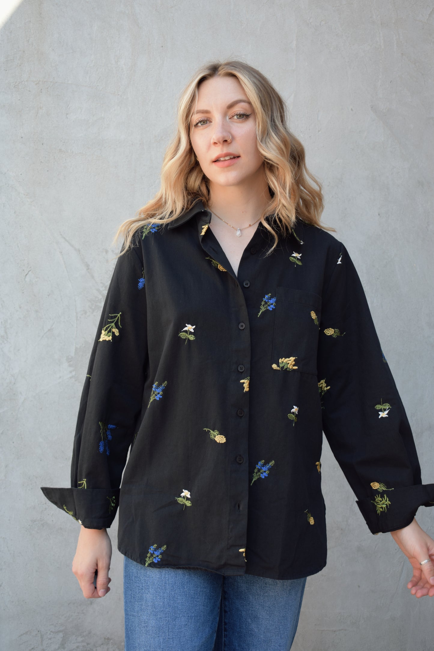 oversized black button down with flowers embroidered on front, back, and sleeves. Flowers are green, blue, yellow, white. Shirt tail hem, buttoned cuffs, one patch breast pockets, collared neckline, black buttons.