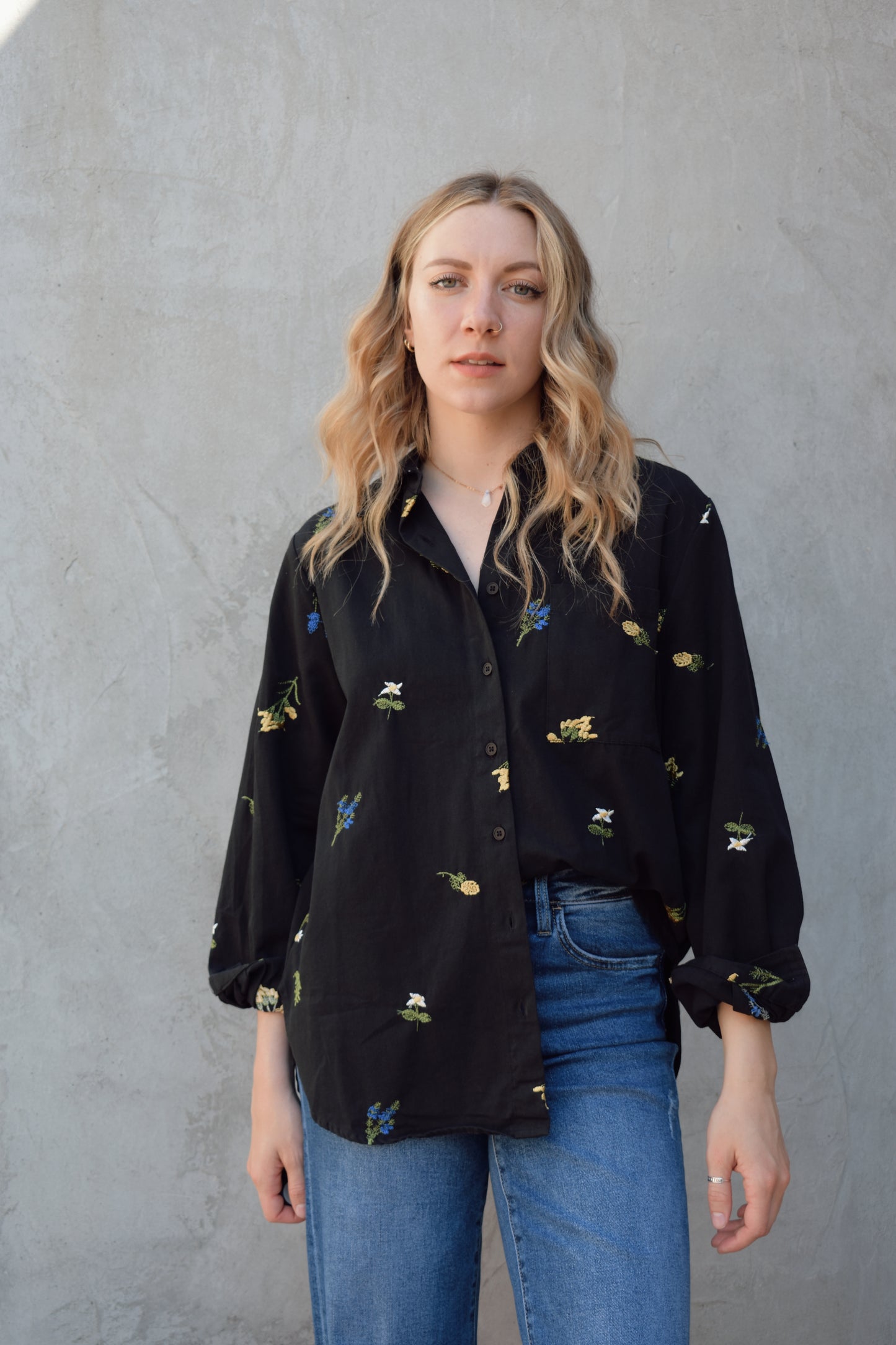 oversized black button down with flowers embroidered on front, back, and sleeves. Flowers are green, blue, yellow, white. Shirt tail hem, buttoned cuffs, one patch breast pockets, collared neckline, black buttons.