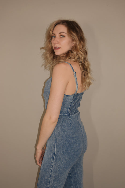 lightly acid washed denim jumpsuit with twisted bodice with a triangle cutout on front. adjustable spaghetti straps. fitted waistband and smocked stretchy back. side zip enclosure. no pockets, flared legs with exposed seam down the front of the legs. stretchy.