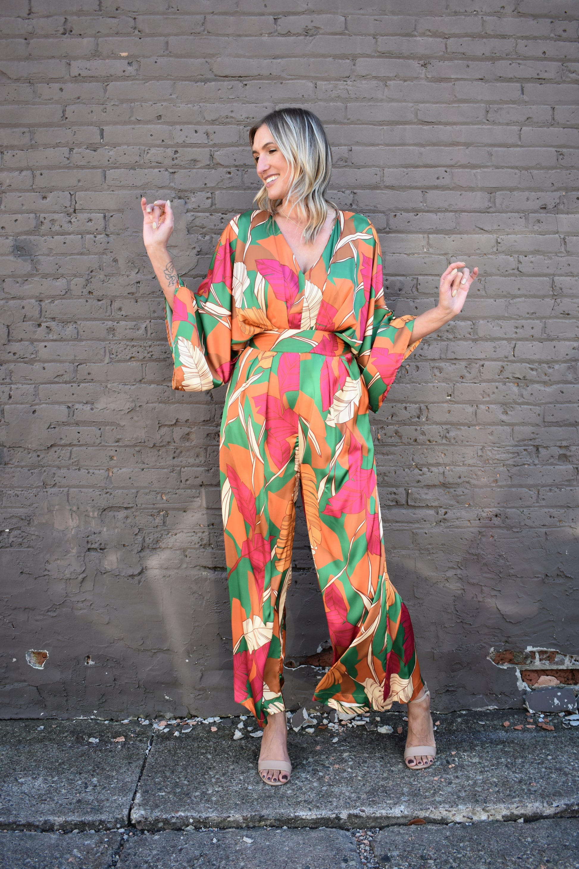 statement satin jumpsuit with a green, rust, cream and pink color scheme. V neckline, kimono sleeves, wide waistband, side pockets, center back slit with button closure on back of neck. wide leg. 