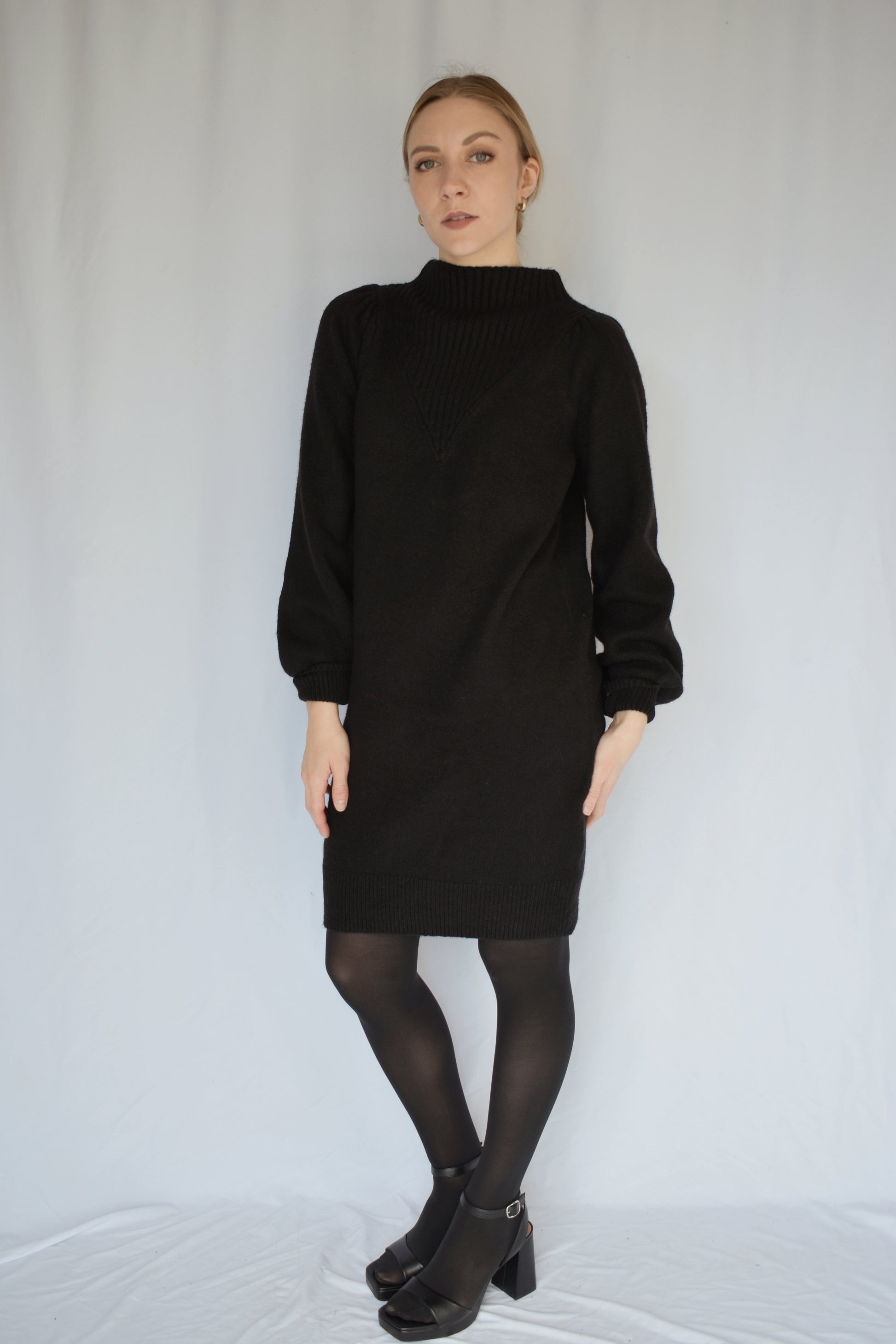 black sweater dress with long sleeves. ribbed cuffs and mock neck with v neck ribbed detail. slight puff on shoulders. hits just above the knees. relaxed fit, not oversized.