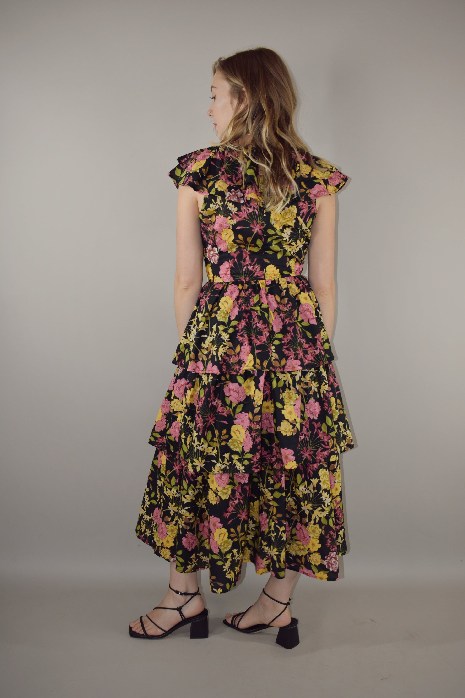 midi tiered floral dress with black background and pink, yellow, green and neutral tones in the floral pattern. v neck with cap flutter sleeves that are also layered. fitted bodice and invisible back zip enclosure. 