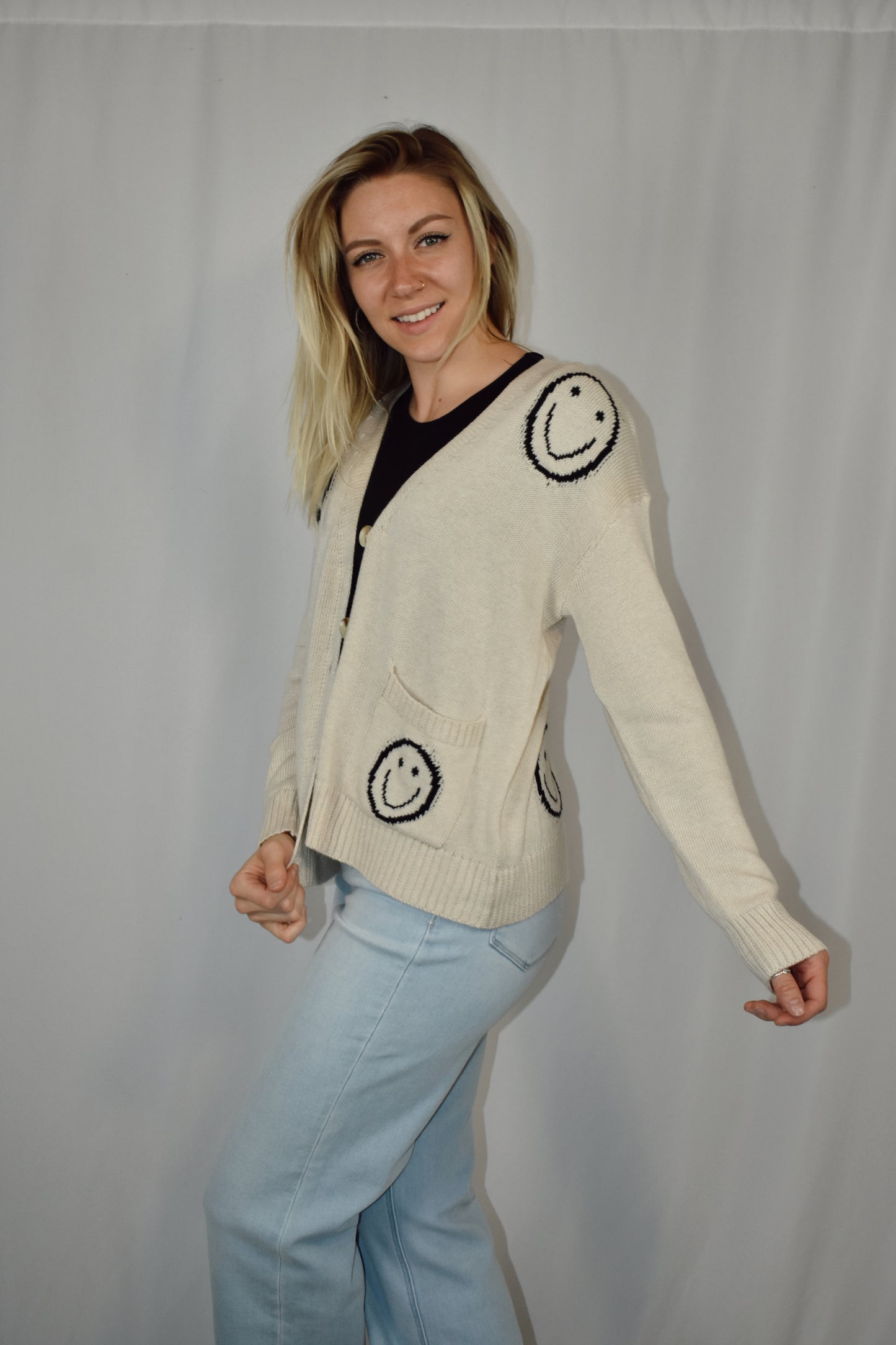 Button down cardigan in a neutral color with big black smiley faces and two patch pockets on the front. Waist length