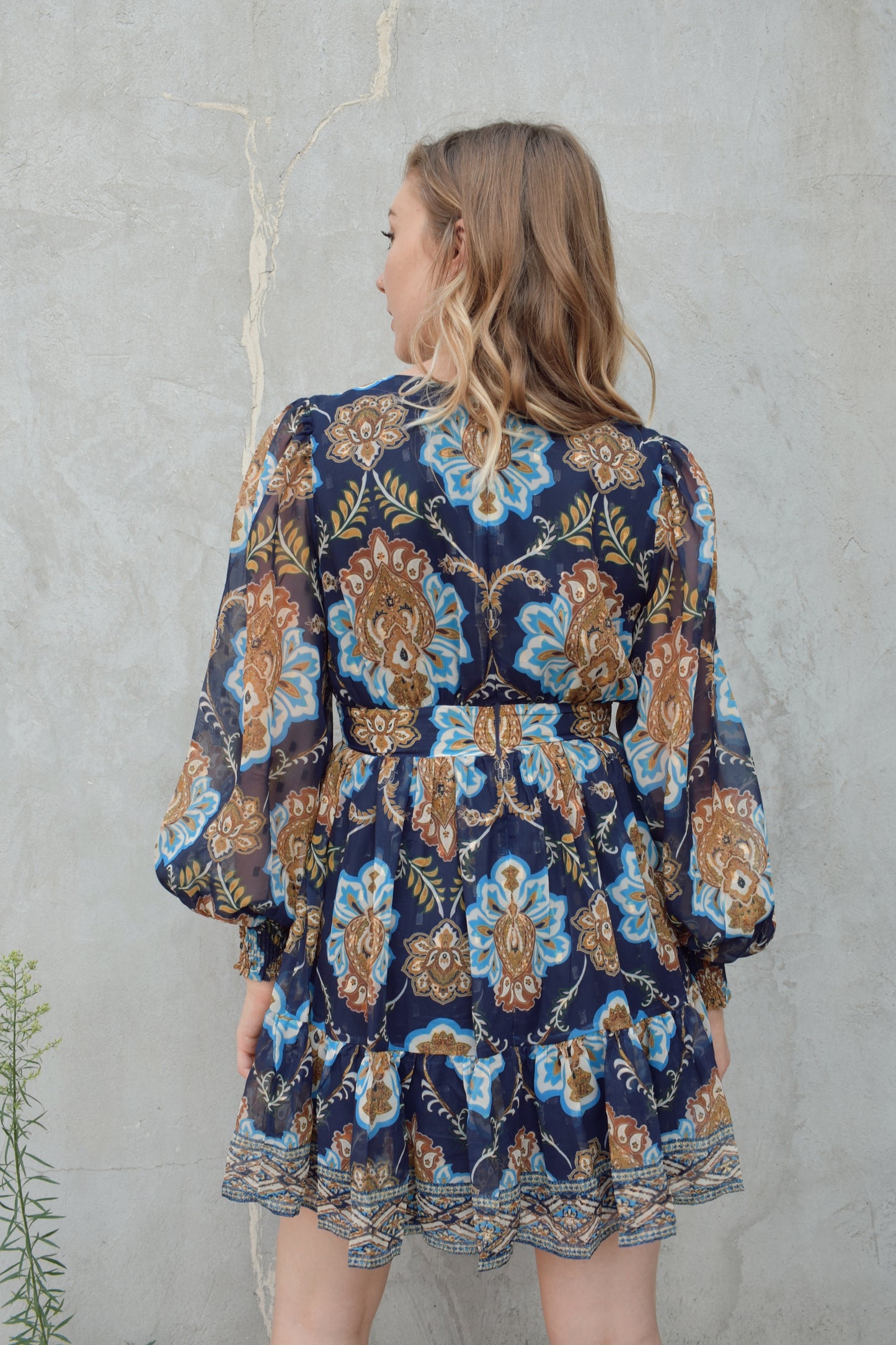 dark blue mini dress with gold, white, and light blue boho pattern. deep v neckline and waist yoke with long sleeves sinched at cuff. back zip enclosure. full skirt. chiffon fabric making sleeves look sheer.