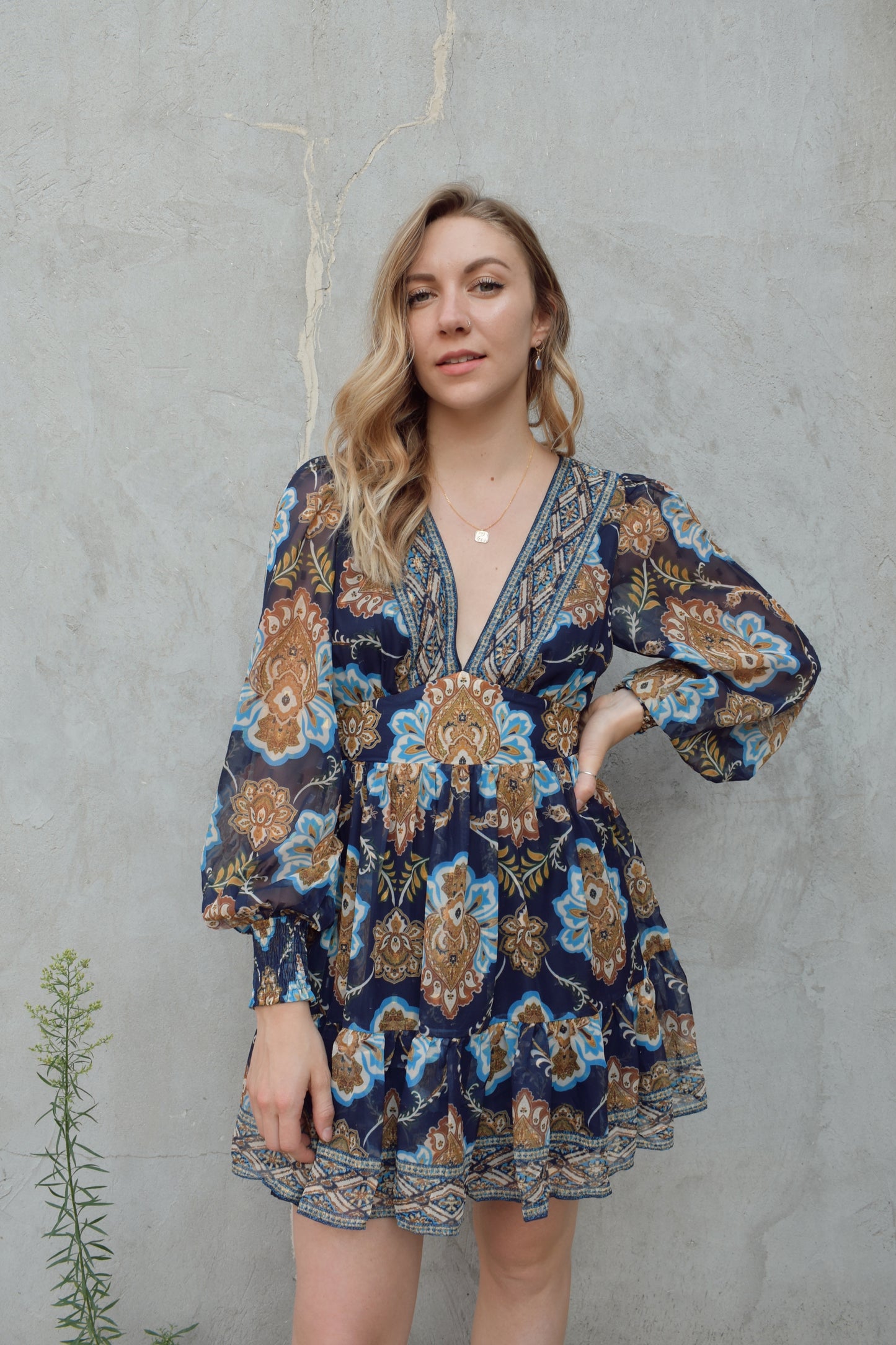 dark blue mini dress with gold, white, and light blue boho pattern. deep v neckline and waist yoke with long sleeves sinched at cuff. back zip enclosure. full skirt. chiffon fabric making sleeves look sheer.